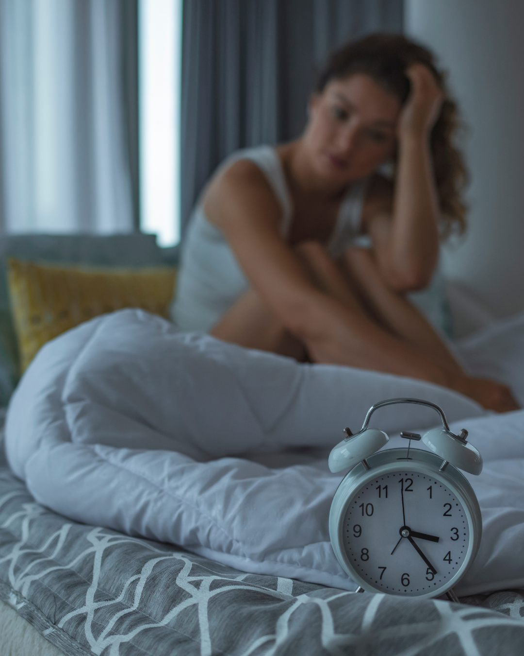 Woman sitting awake in bed looking tired beside alarm clock, representing insomnia, poor sleep and disrupted sleep patterns