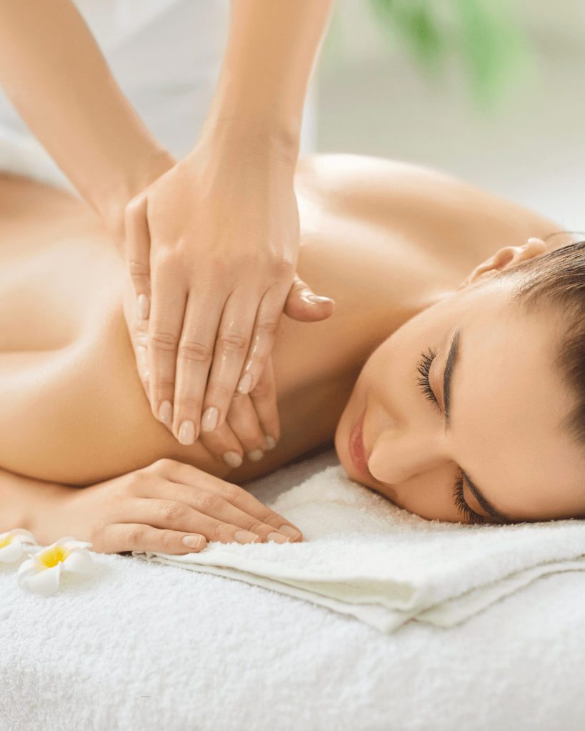 A woman receiving a relaxing massage on a treatment table, with her eyes closed and a towel beneath her. A hand is gently applying pressure on her back, and a flower is placed on the table.