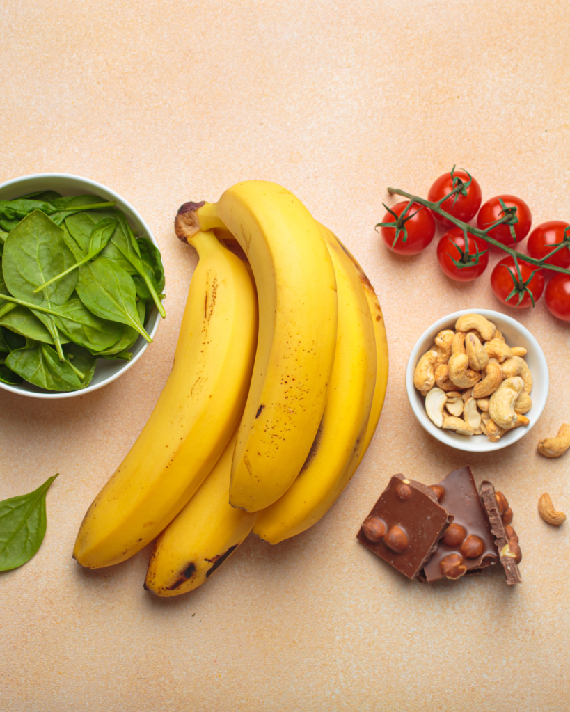 A flat lay of fresh bananas, a bowl of spinach, cherry tomatoes on the vine, a bowl of cashews, and a piece of chocolate with nuts on a beige background.