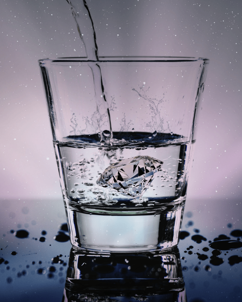 A glass filled with water and a diamond submerged underneath, with water splashing around and a reflective surface.