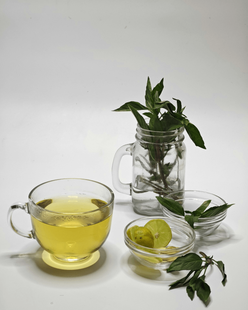 A glass cup of herbal tea with a bright yellow color, accompanied by a jar with fresh herbs and bowls containing lime and green leaves.