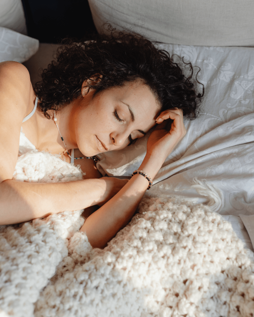 A woman with curly hair lying on a soft bed, covering herself with a fluffy blanket, appearing peaceful and relaxed.