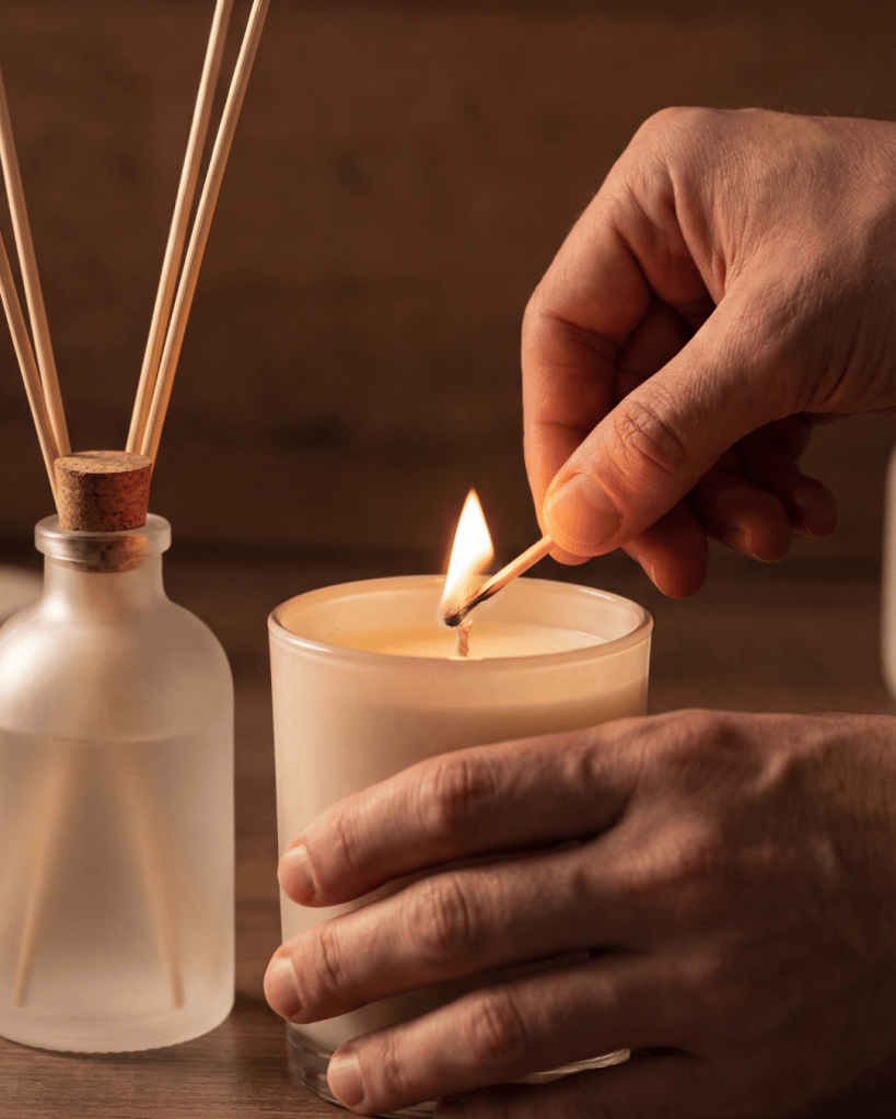 A hand lighting a candle with a matchstick, next to a decorative glass bottle with reed sticks in it.
