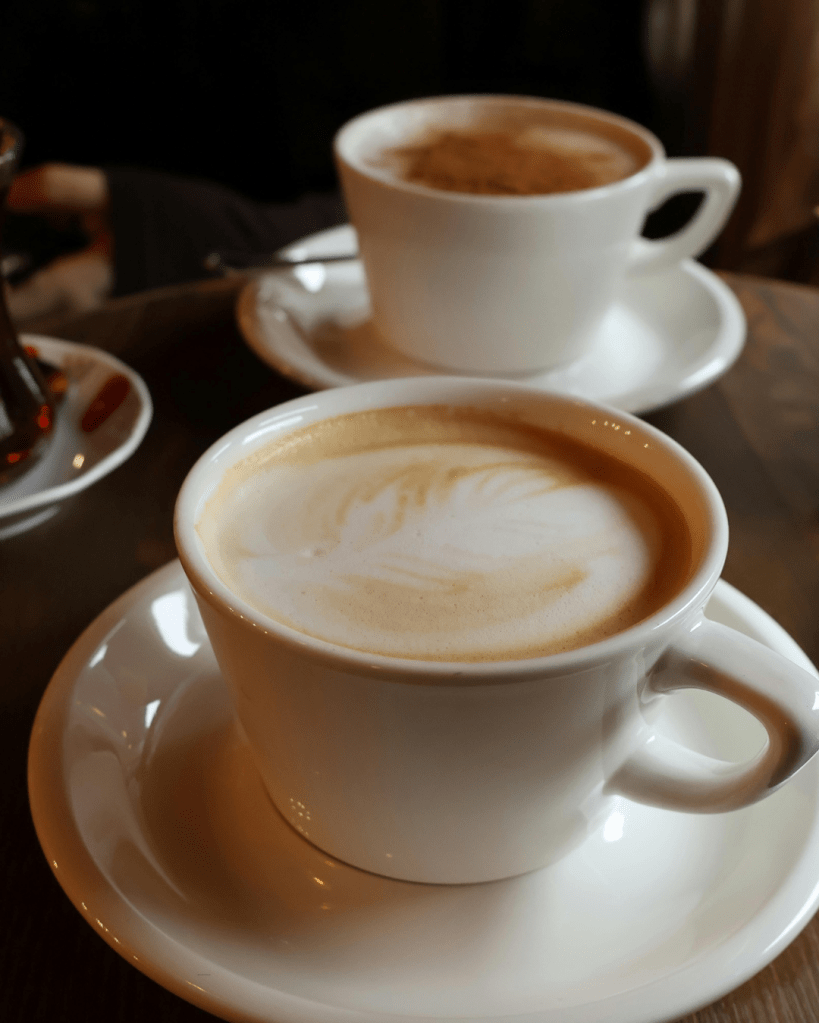 A close-up image of two white cups of coffee on saucers, one in the foreground with a light-colored latte art, and another in the background.