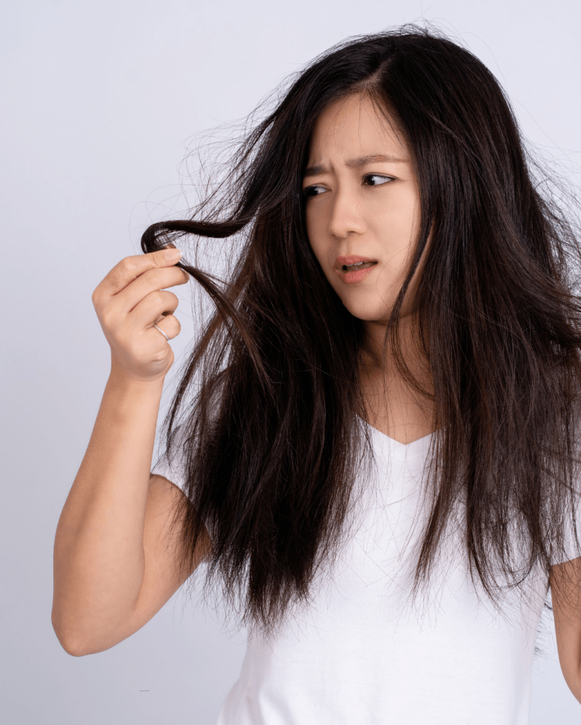 A woman with long, frizzy hair looks concerned while holding a strand of her hair, wearing a plain white t-shirt.
