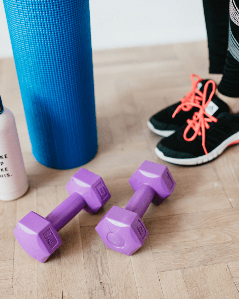 A pair of purple dumbbells on a wooden floor, next to a blue yoga mat and a white water bottle, with a person wearing black sneakers and bright orange laces nearby.