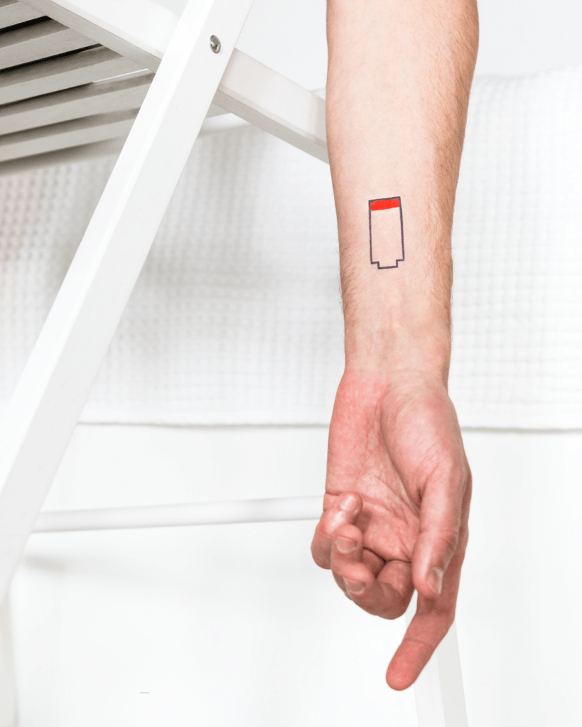 A close-up of a hand showing a tattoo of a battery icon with a red top on the forearm.