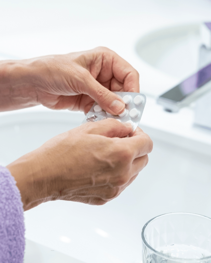 A person holding a blister pack of white tablets, preparing to remove a pill, with a glass of water in the foreground.