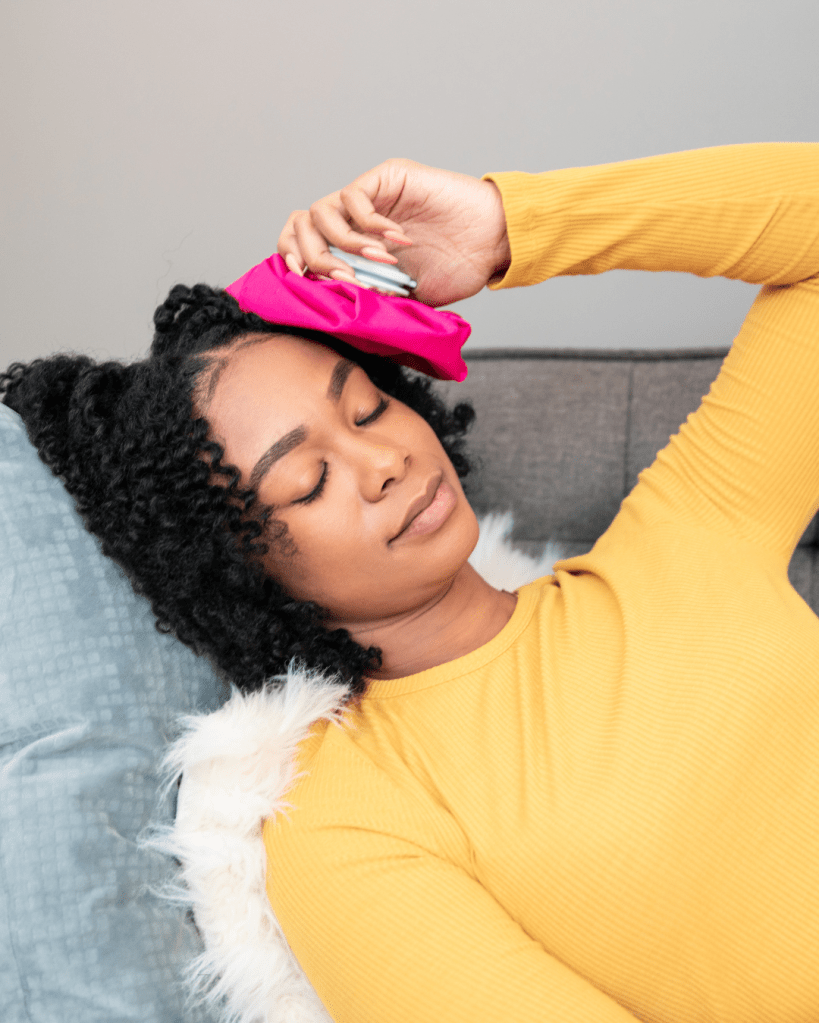 A woman with curly hair relaxing on a couch, holding a pink heat pack to her forehead, wearing a yellow long-sleeve shirt.