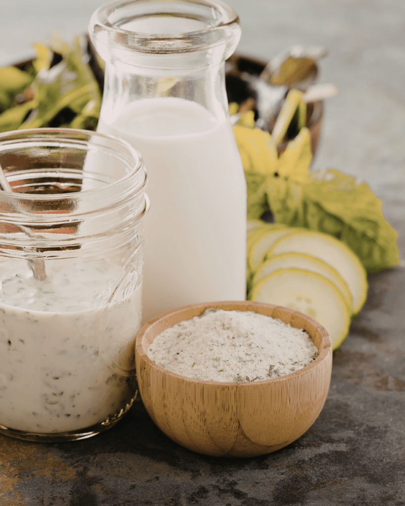 A mason jar filled with creamy dressing, a glass bottle of milk, and a wooden bowl of seasoning mix, with fresh ingredients in the background.