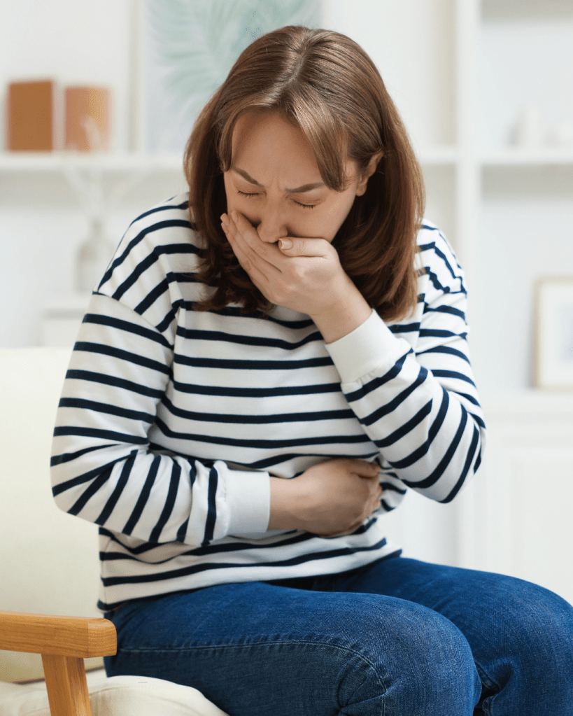 A woman sitting on a chair, holding her stomach and covering her mouth, showing signs of discomfort or nausea.