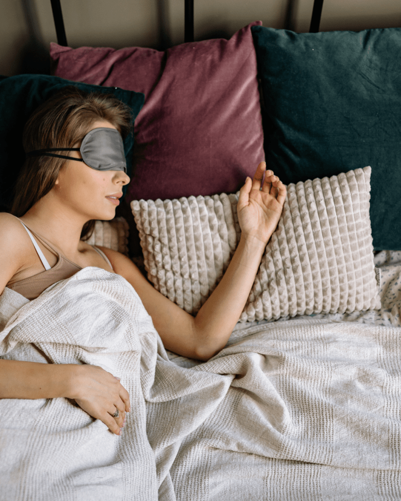 A woman resting in bed wearing a sleep mask, with her arm extended over a textured pillow while covered with a light blanket. Surrounding her are colorful pillows.