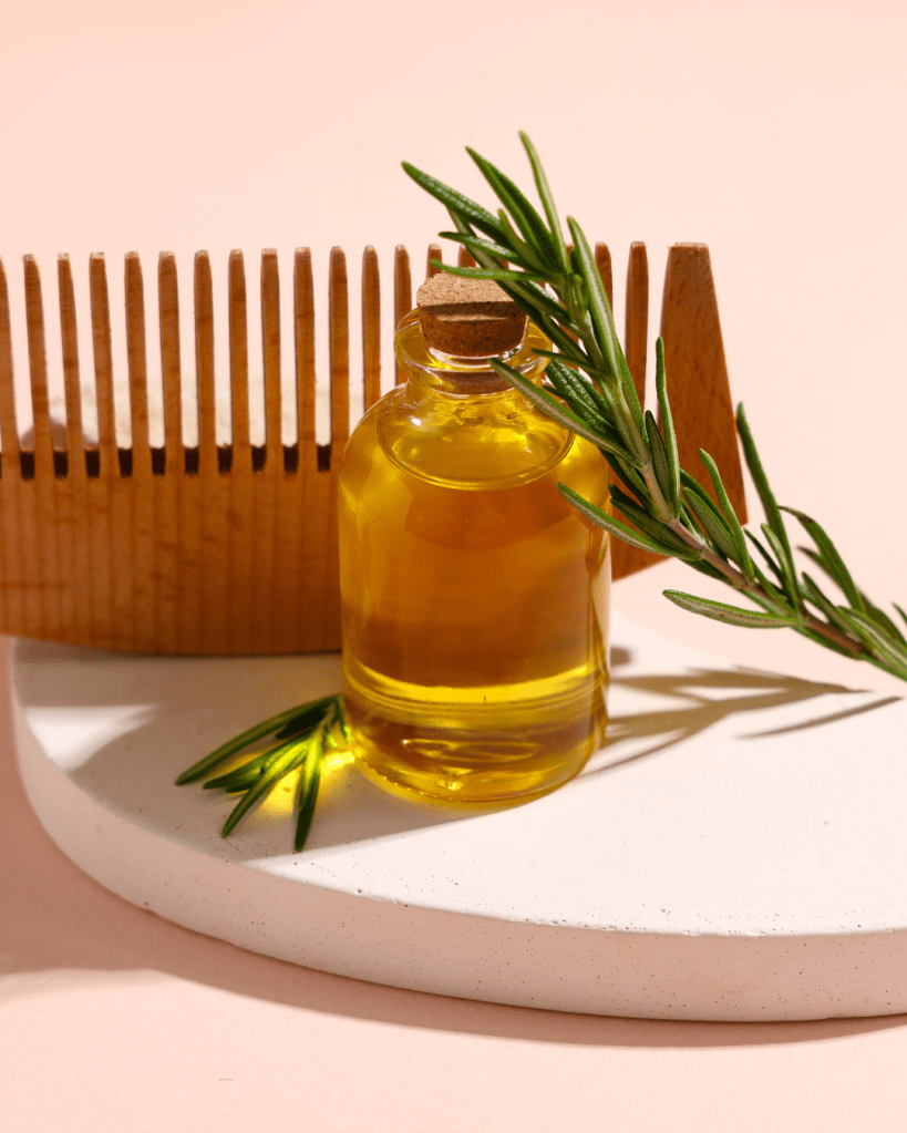 A glass bottle of golden oil with a cork lid, placed on a white circular surface alongside a wooden comb and a sprig of fresh rosemary.