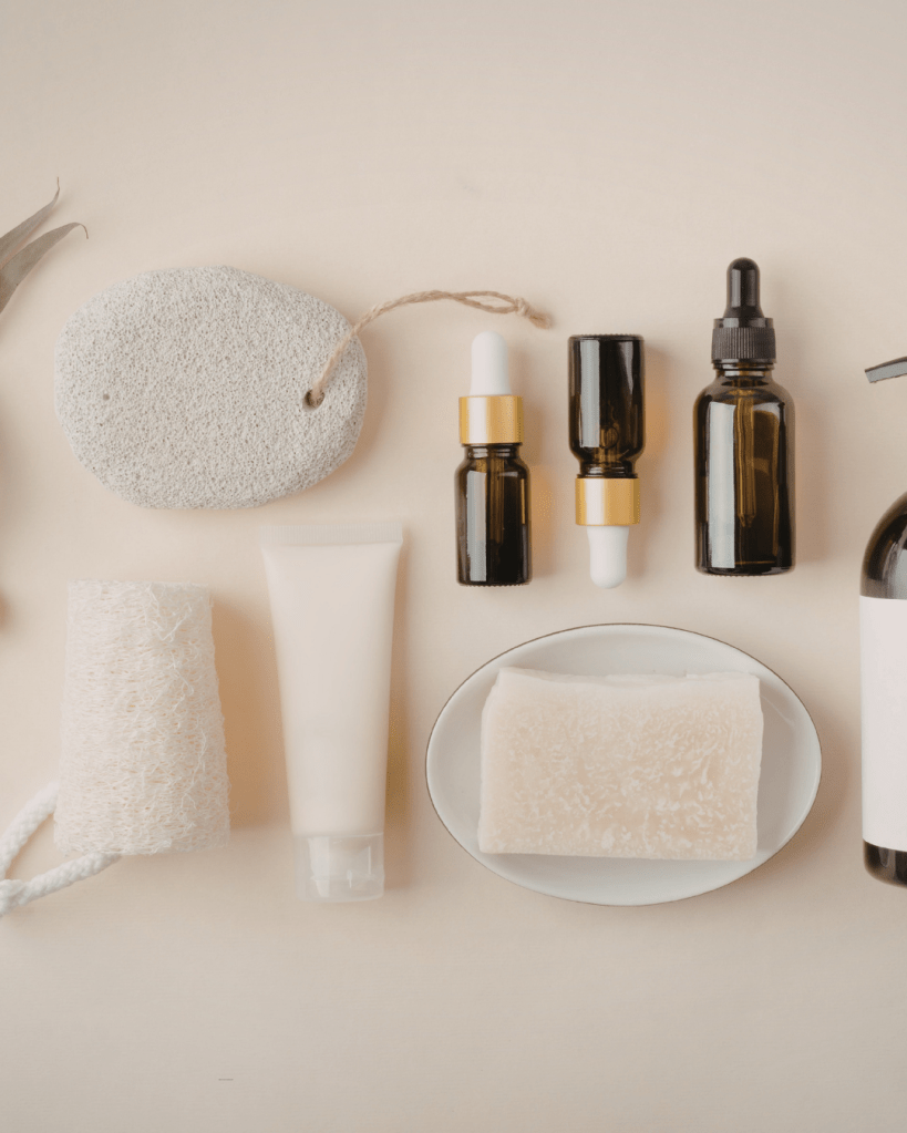 Various skincare products arranged on a light surface, including essential oils in dark bottles, a white cream tube, a soap bar on a plate, a loofah, and a pumice stone.