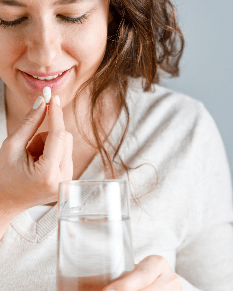 A woman holding a white pill above her lips with a glass of water in her other hand, preparing to take the medication.