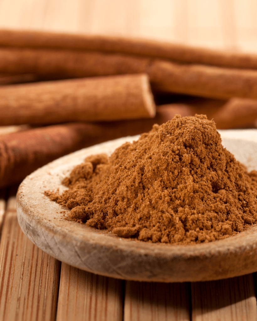 A close-up of a mound of ground cinnamon powder in a small dish, with whole cinnamon sticks in the background on a wooden surface.