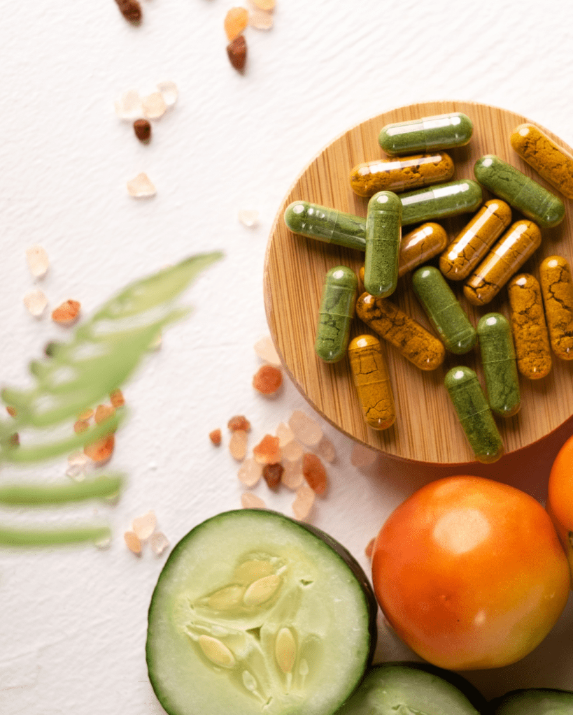 A wooden bowl filled with colorful dietary supplements, surrounded by sliced cucumber, tomatoes, and decorative salt grains on a light background.