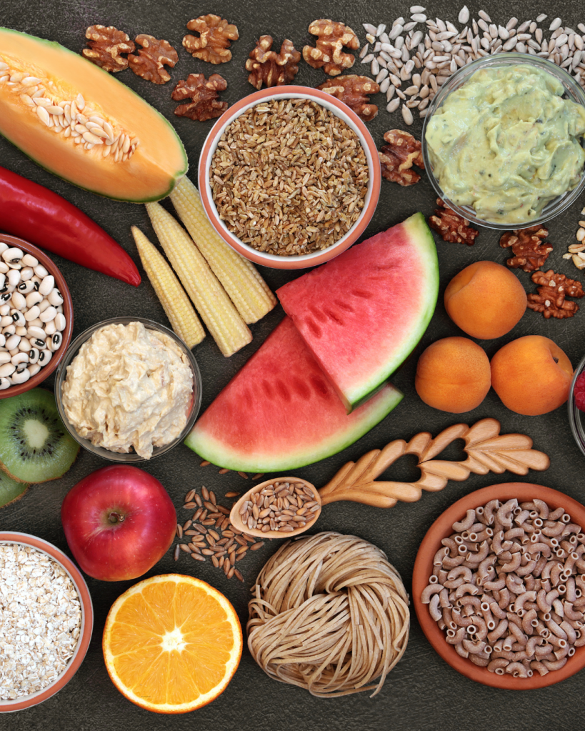 A variety of colorful fruits, grains, and nuts arranged on a dark surface, including watermelon, cantaloupe, an apple, an orange, apricots, and kiwi, alongside bowls of seeds, pasta, and oatmeal.