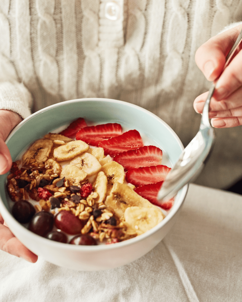 A bowl of yogurt topped with bananas, strawberries, granola, and dark chocolate chips, held by a person with a spoon ready to eat.
