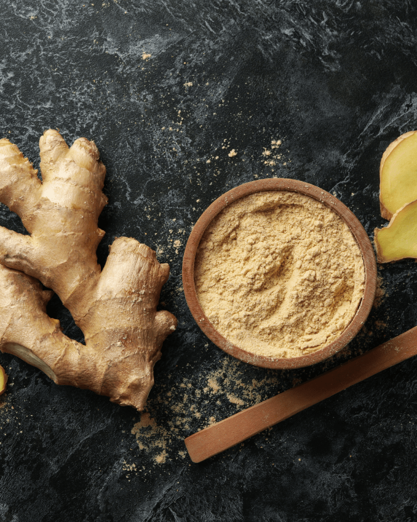 Fresh ginger root alongside a bowl of ginger powder and a piece of sliced ginger on a dark textured surface.