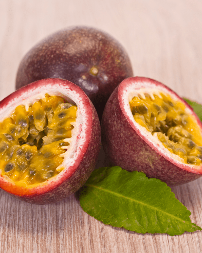 Three passion fruits, two cut in half to reveal their vibrant yellow pulp and seeds, resting on a wooden surface with a green leaf.