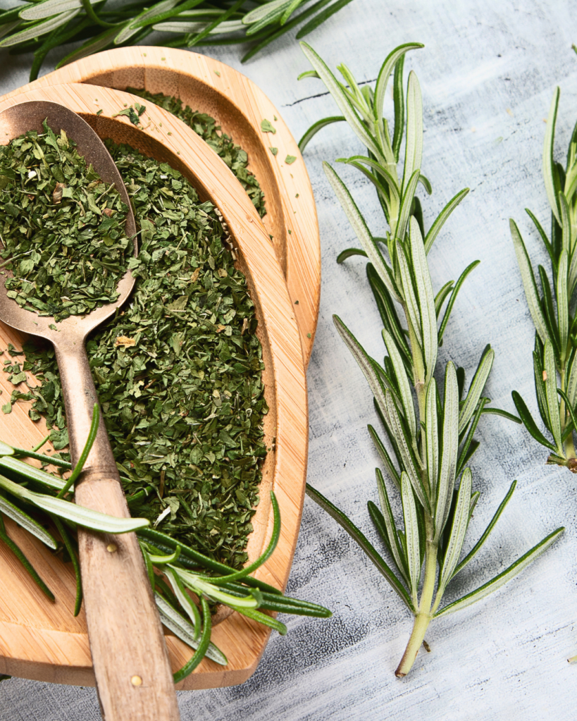 Fresh rosemary sprigs and dried rosemary leaves in wooden bowls with a spoon on a textured surface.