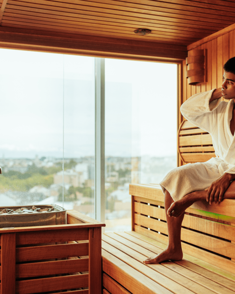 A person sitting in a wooden sauna, wearing a white bathrobe, while looking thoughtfully out of a large glass window with a cityscape in the background.