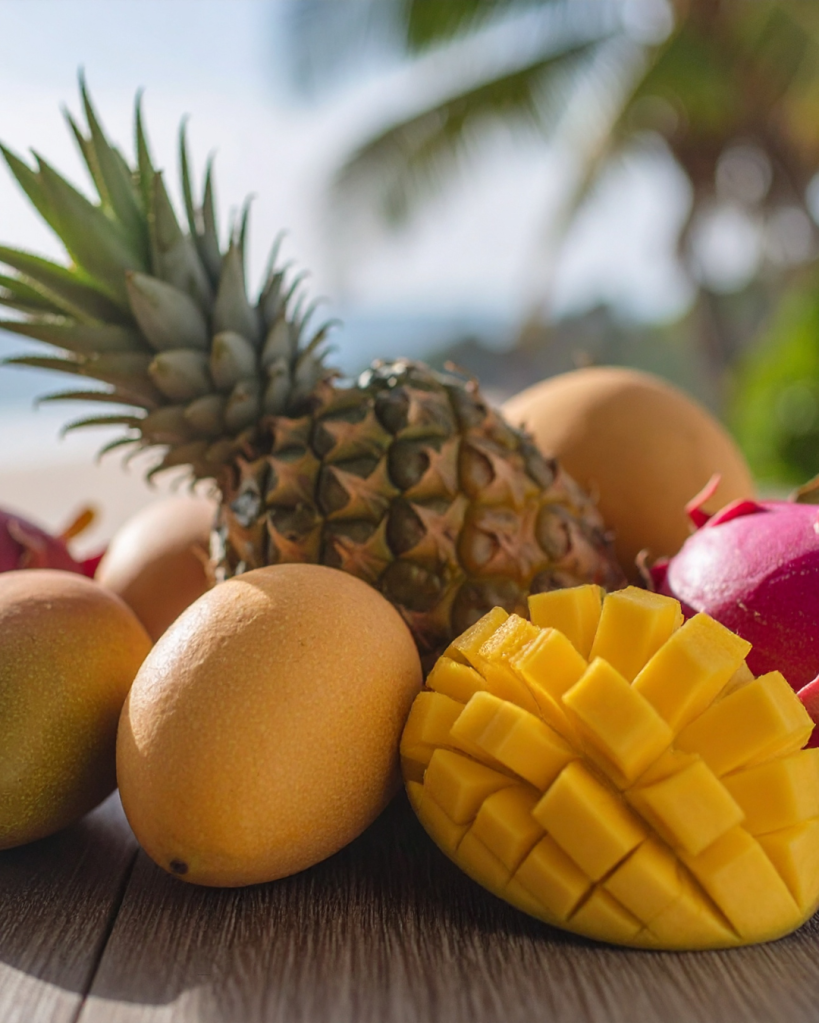 A colorful arrangement of tropical fruits, including a pineapple, mango, and dragon fruit, set against a blurred beach background.