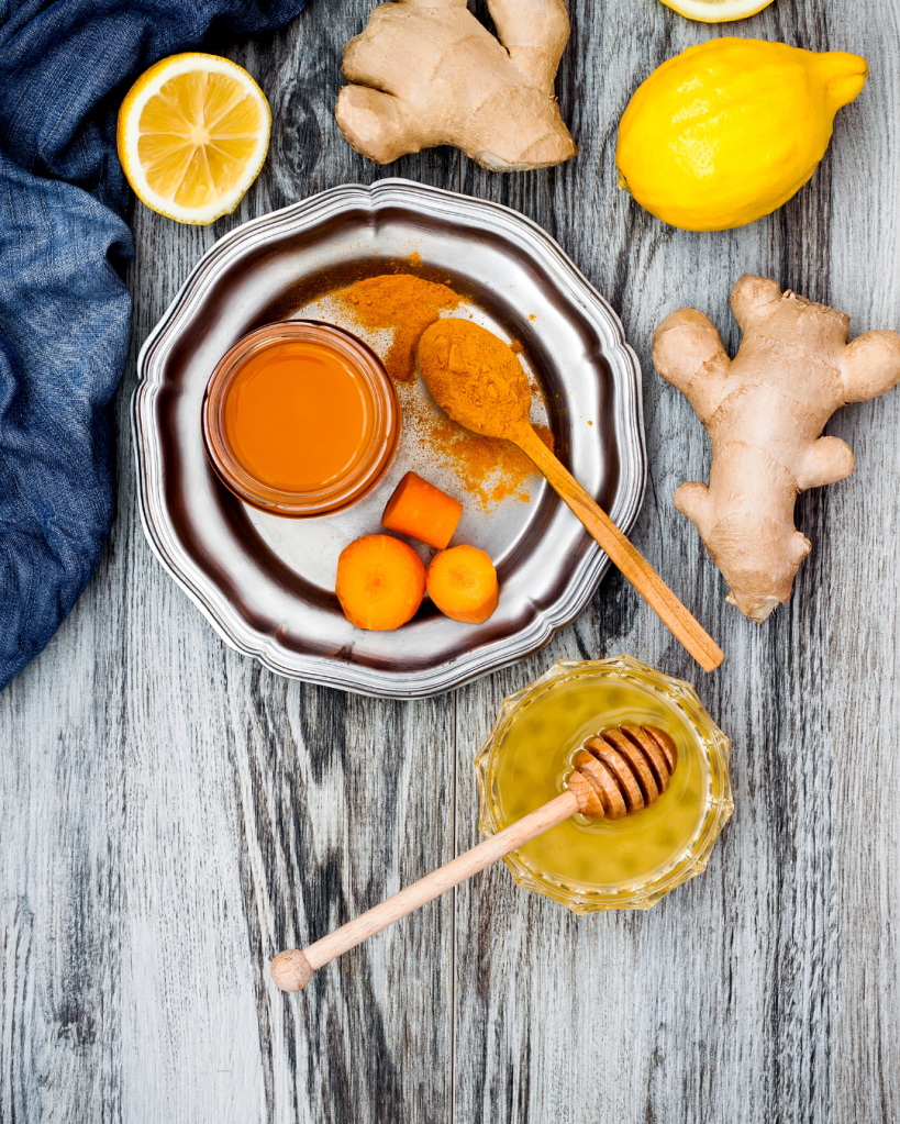 A collection of fresh ingredients including ginger, lemon, and carrots arranged around honey and a jar of turmeric on a silver plate, set against a wooden background.