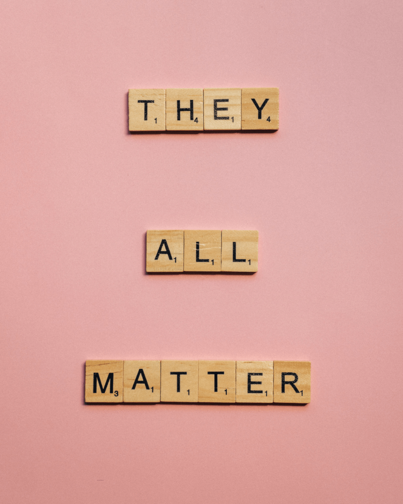 Scrabble tiles forming the phrase 'They All Matter' against a pink background.