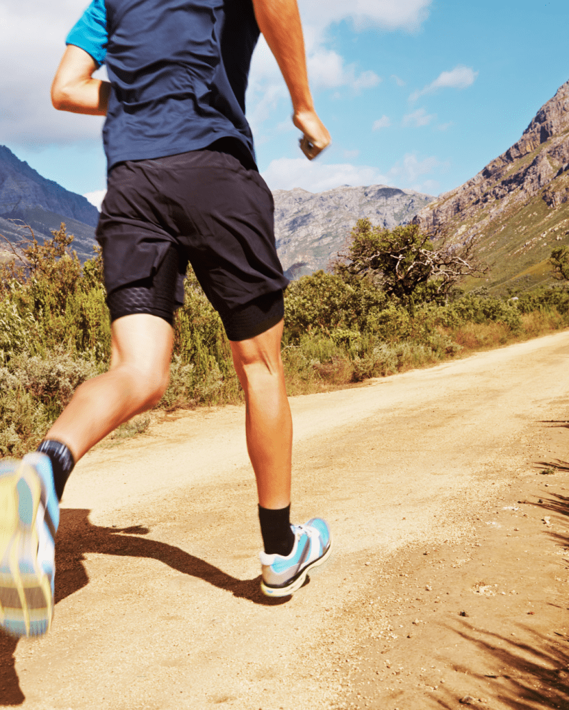 A person running on a dirt path with mountains in the background, wearing athletic clothing and running shoes.