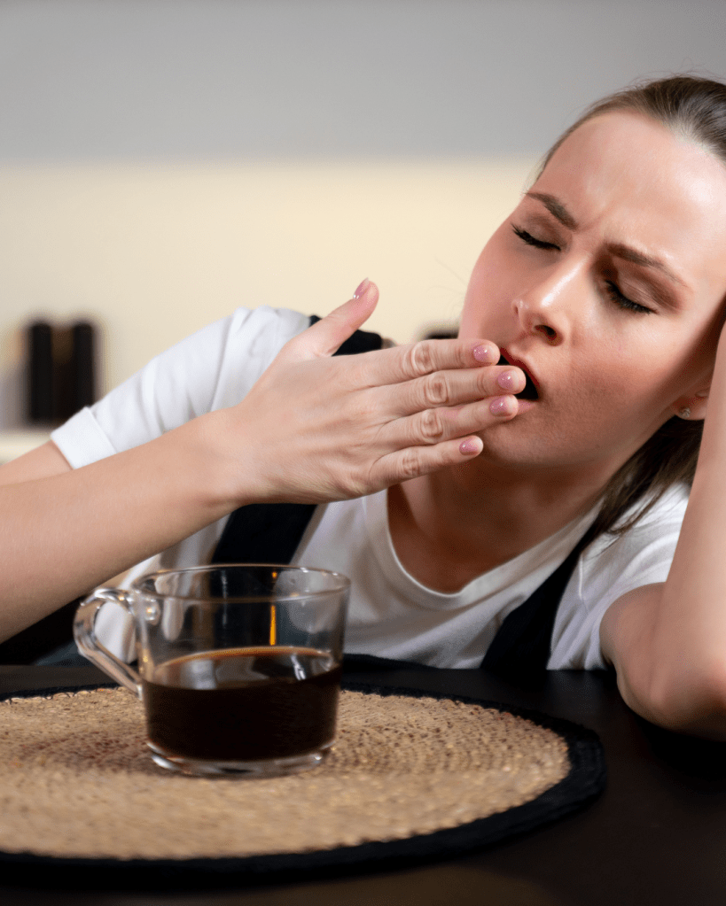 A woman yawning while sitting at a table with a cup of coffee in front of her, looking tired.