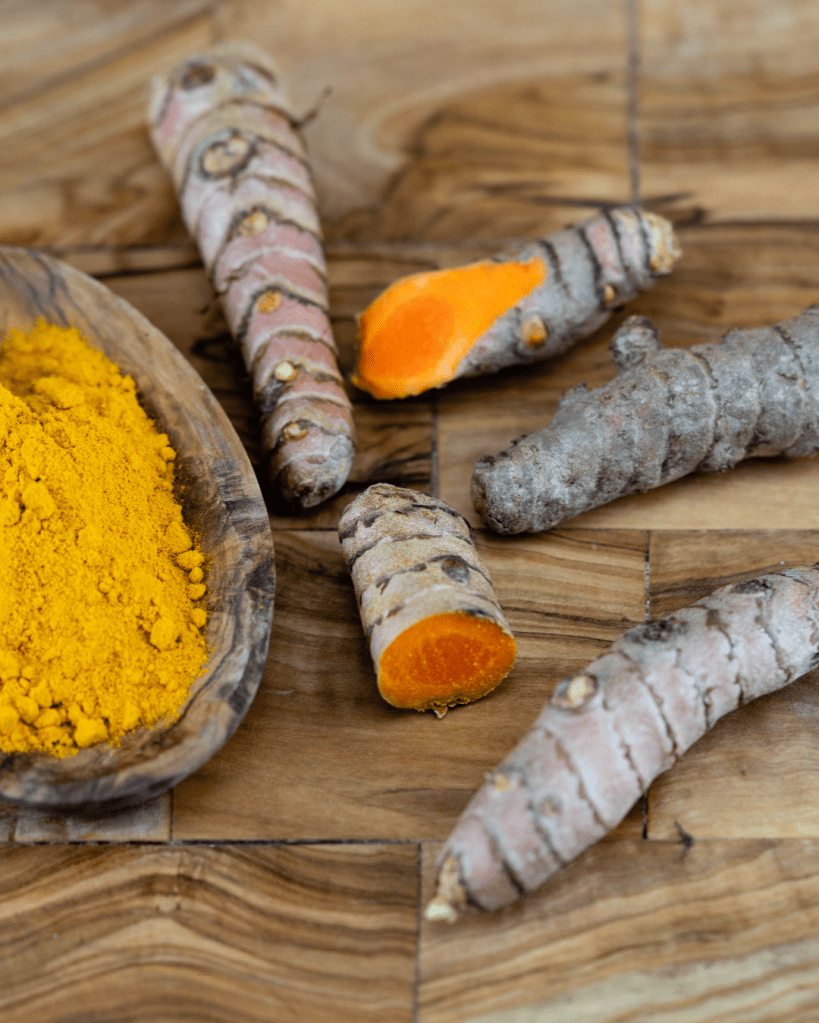 A wooden bowl filled with bright yellow turmeric powder, surrounded by fresh turmeric roots, some of which are cut open to reveal their orange interior, placed on a wooden surface.