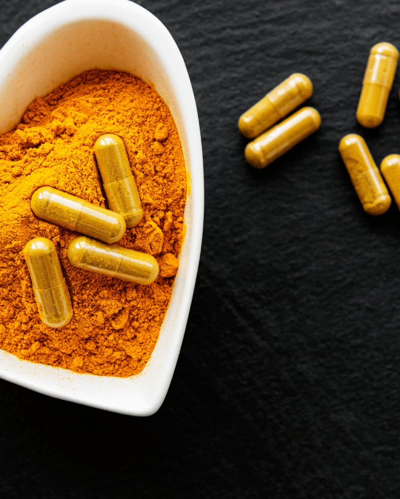 Turmeric powder in a heart-shaped dish with yellow capsules beside it on a dark surface.