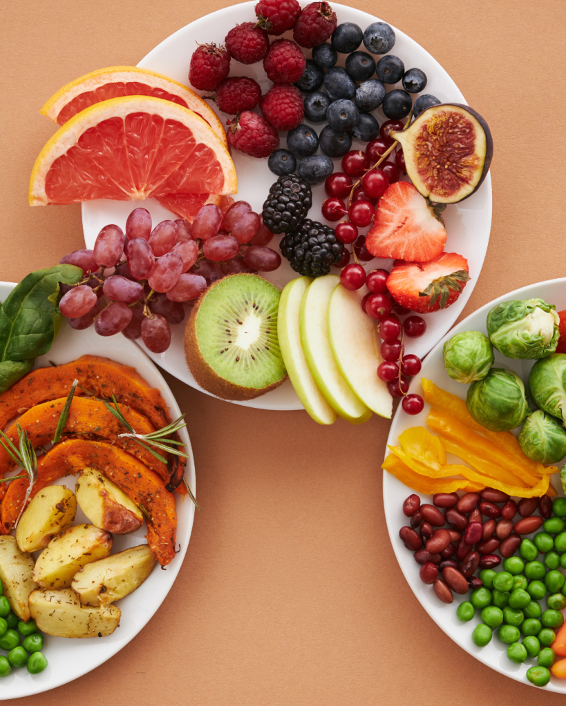 Three plates of colorful fruits and vegetables, including sliced grapefruit, berries, grapes, kiwi, apple, roasted pumpkin, potatoes, green peas, and brussels sprouts, arranged aesthetically on a brown background.
