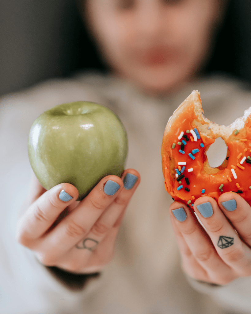 A person holds a green apple in one hand and a half-eaten orange donut with colorful sprinkles in the other, showcasing a choice between healthy and indulgent food.