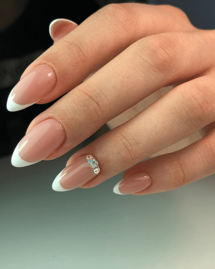 Close-up of a hand displaying elegant acrylic nails with a nude base and white French tips, featuring a small gemstone accent on the ring finger.