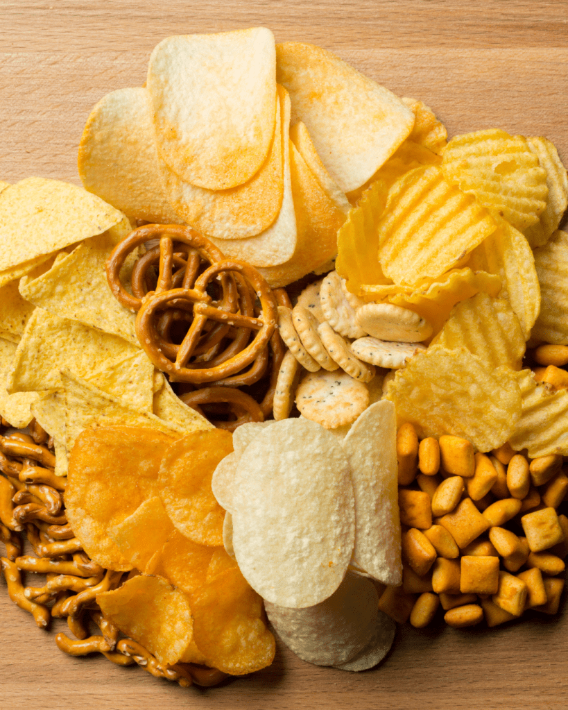 A variety of snacks arranged on a wooden surface, including potato chips, pretzels, crackers, and cheese puff snacks.