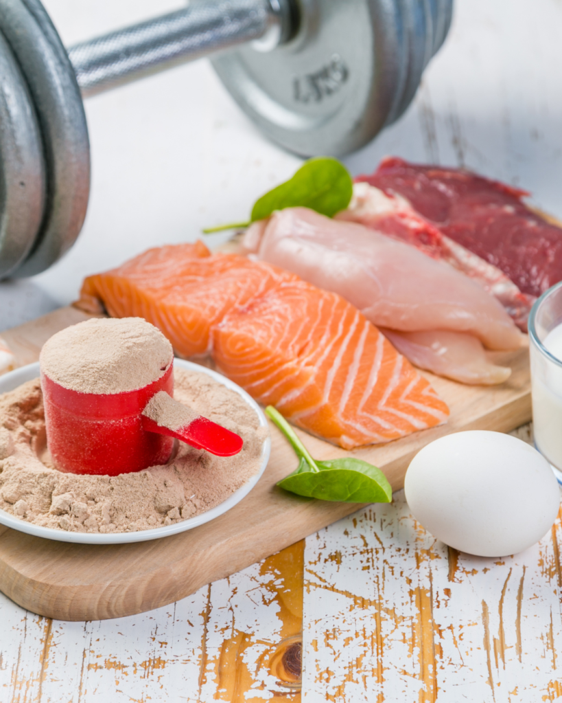 A variety of protein sources including salmon, chicken, and beef, accompanied by a red measuring cup filled with protein powder, a raw egg, spinach leaves, and a glass of liquid on a wooden cutting board, with a dumbbell in the background.