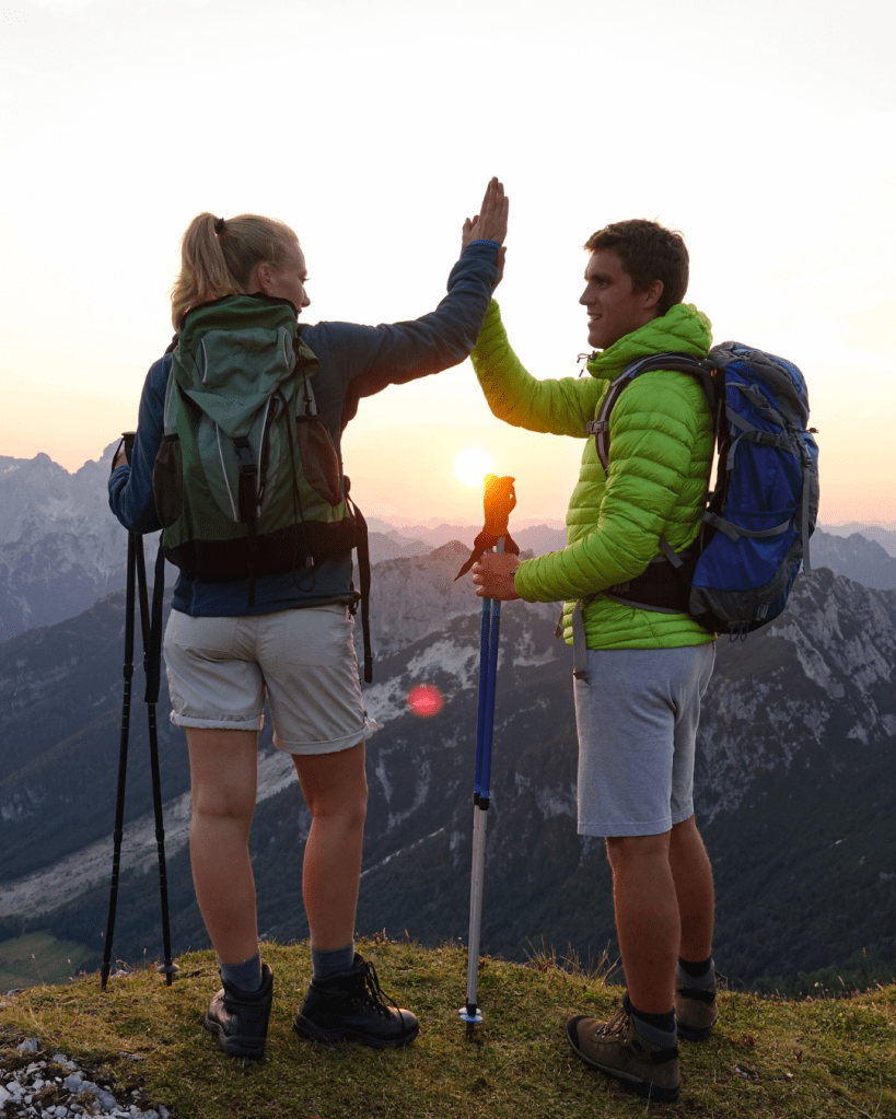 Two hikers high-fiving on a mountain top at sunset, surrounded by scenic mountain views.
