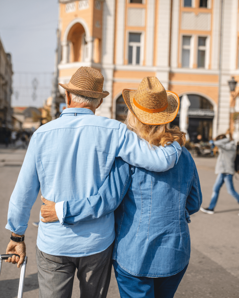 An elderly couple walking together, embracing affectionately while wearing straw hats, with a city street and historic building in the background.