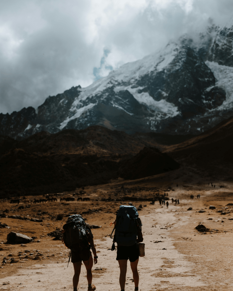 Two hikers with backpacks walking along a dusty trail in a mountainous landscape with dramatic clouds and snow-capped peaks in the background.