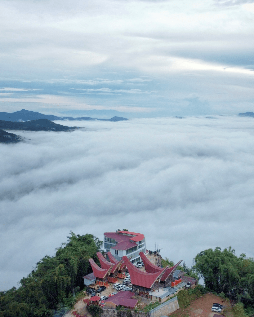 Aerial view of a building complex with unique architecture, surrounded by a sea of clouds and rolling hills under a cloudy sky.