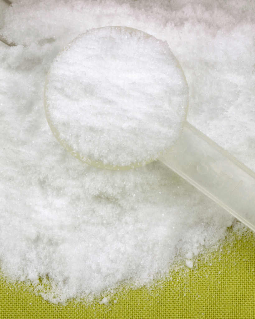 A close-up image of a measuring spoon filled with white granulated sugar on a textured green surface.