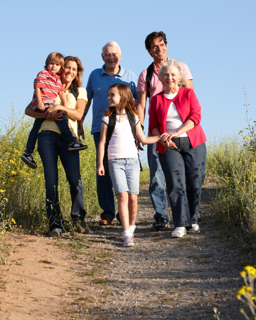 A happy family of five walking together on a dirt path, surrounded by greenery and yellow wildflowers on a sunny day.