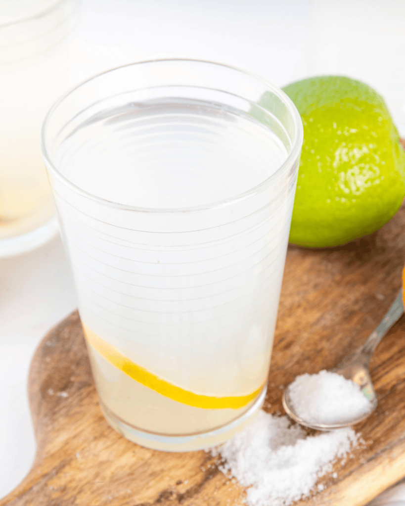 A clear glass of lemonade with a slice of lemon inside, placed on a wooden surface next to a green lime and a teaspoon of salt.