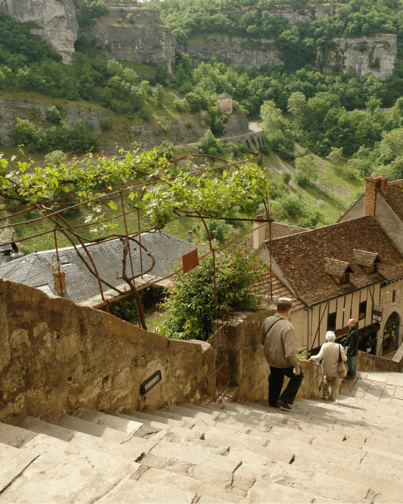 An elderly couple walking down stone steps in a lush green landscape, surrounded by trees and historic buildings.