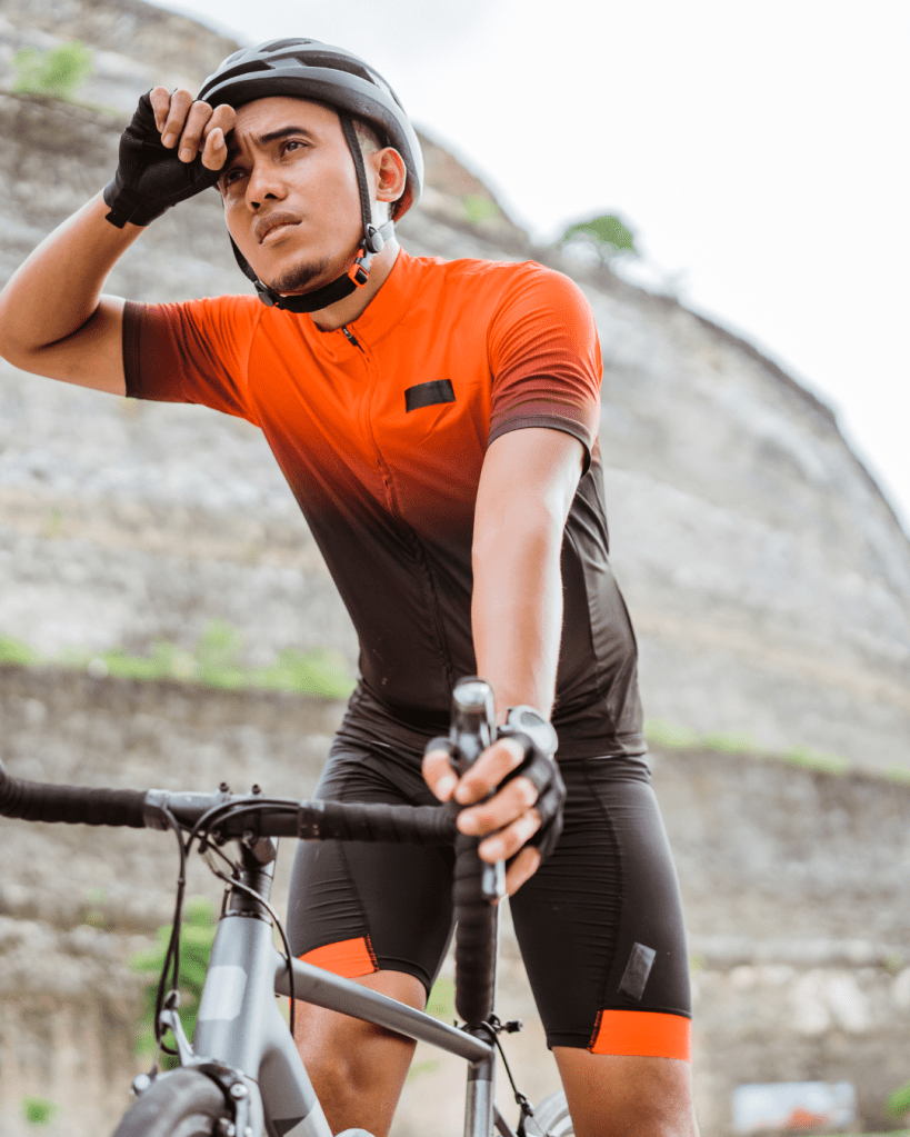 A cyclist in an orange and black cycling outfit wipes sweat from his forehead while standing next to his bike, with a rocky landscape in the background.