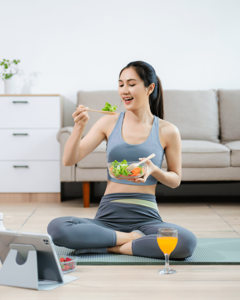 A woman sitting cross-legged on a yoga mat, enjoying a salad from a bowl while smiling. She is dressed in workout attire, with a glass of orange juice beside her and a tablet on the mat.