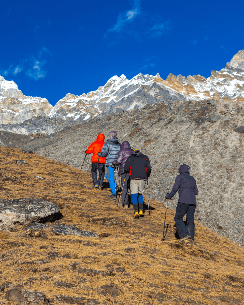 Group of hikers trekking up a mountain trail with snow-capped peaks in the background, wearing colorful jackets.